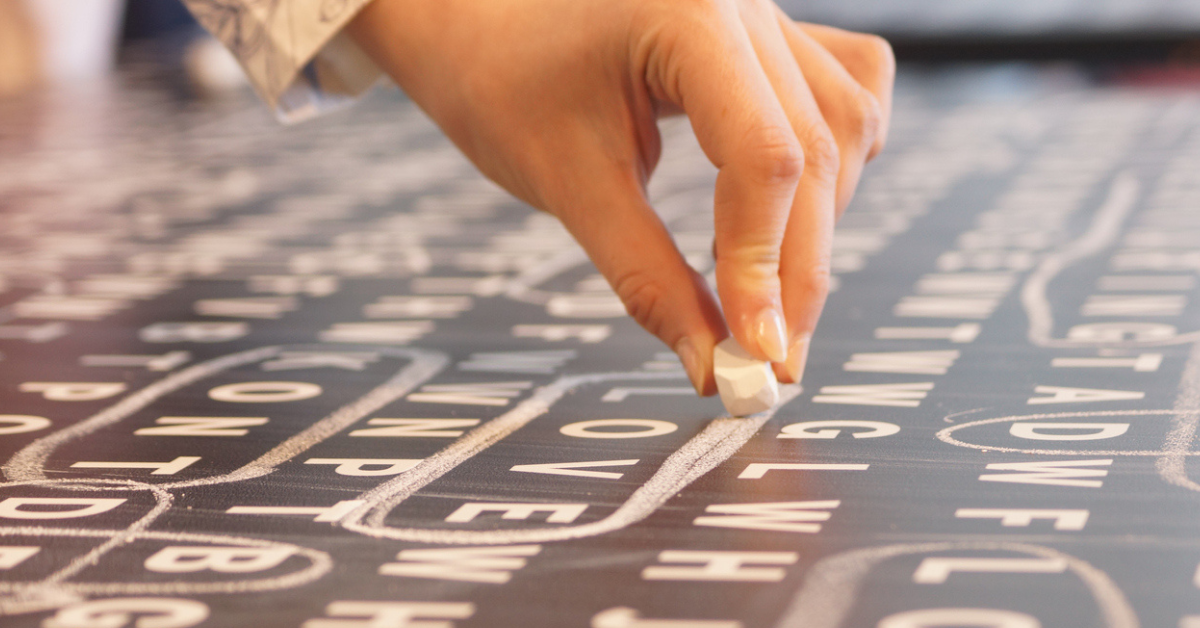 Delicate woman's hand encircling a word with white chalk on black board filled with letters.